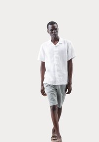 Weiße Leinenkurzarmbluse und hellgraue Shorts, getragen von einem Model. Sandalen und minimale Details ergänzen das lässige Outfit.
