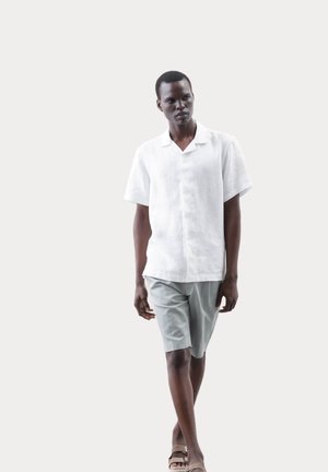 Camicia bianca in lino a maniche corte e pantaloni corti grigio chiaro indossati da un modello. Sandali e dettagli minimal completi il look casual.