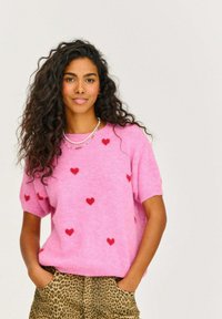 Ružový pletený sveter s krátkymi rukávmi, ozdobený malými červenými srdiečkami. V kombinácii s nohavicami s leopardím vzorom, ruky v vreckách.