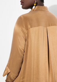 Camicia in satin dorato con colletto, caratterizzata da un motivo testurizzato e una piega centrale sul retro. Maniche a tre quarti con fessure laterali.
