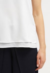 Débardeur blanc sans manches en tissu lisse, mettant en avant un design superposé avec un ourlet légèrement arrondi. Main visible au bord, montrant la texture satinée.