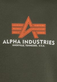 Camiseta de color verde oliva con un logotipo texturizado en naranja de la letra "A" y el texto "ALPHA INDUSTRIES KNOXVILLE, TENNESSEE, U.S.A." en blanco.