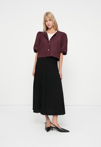 Leichte, burgunderrote, cropped Jacke mit strukturiertem Muster und kurzen Puffärmeln, kombiniert mit einem langen schwarzen, plissierten Rock und schwarzen spitzen Pumps.