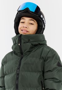Grøn isoleret pufferjakke med høj krave, frontlynlås og logodetalje. Båret med en sort skihjelm og reflekterende blå skibriller.
