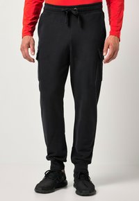 Muž nosící černé cargo tepláky se stahovací šňůrkou v pase a černé tenisky, zobrazený od pasu k nohám na jednobarevném pozadí.