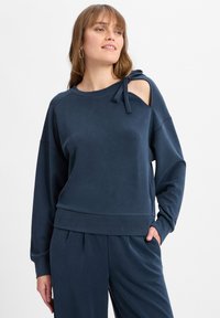 Dunkelblauer Pullover mit lockerer Passform, Schulterausschnitt und Schleifenakzent. Weiche Textur und gerippter Saum für zusätzlichen Komfort.