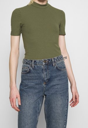 Persoon draagt een geribbelde, olijfgroene top met korte mouwen, ingestopt in een hooggetailleerde, verwassen blauwe spijkerbroek, met een zichtbare tatoeage op de linker onderarm.