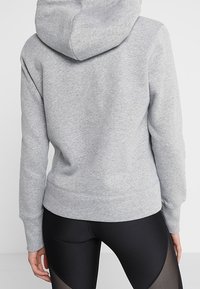 Vrouw in een lichtgrijze hoodie en zwarte leggings met meshpanelen, weergegeven vanuit de rug tegen een effen achtergrond.