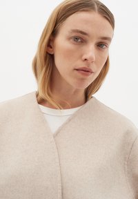 Bežinis vilnonis apsiaustas su minkšta tekstūra ir V formos dekoltė, dėvimas ant balto ilgomis rankovėmis marškinio, rodoma prieš vientisą foną.