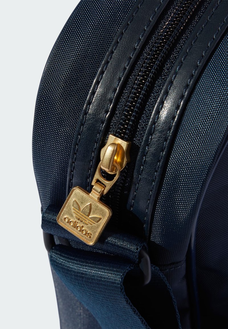 Navy Tasche aus strukturiertem Material, glänzenden Akzenten und einem goldenen Reißverschluss, mit einem quadratischen Anhänger, der das Adidas-Logo zeigt.