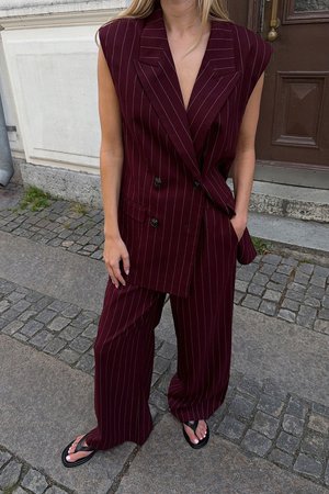 Blazer bordeaux à rayures, avec un design croisé, associé à un pantalon large assorti. Des tongs noires complètent la tenue.