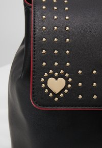 Rabattre de sac en cuir noir avec de petits clous dorés formant une forme de cœur et un motif de points dispersés, bordé d'un fin liseré rouge.