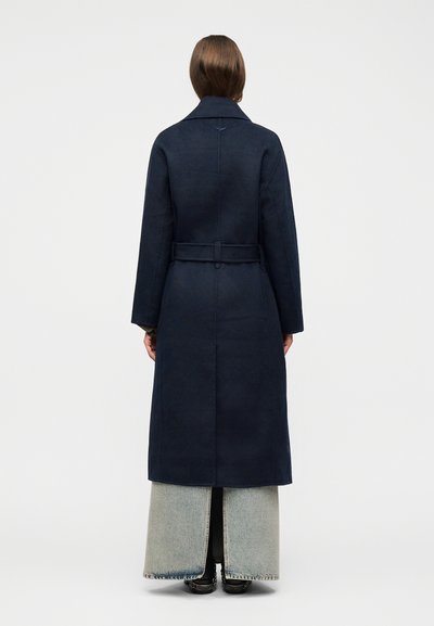 Άτομο που φοράει μακρύ σκούρο ναυτικό παλτό με ζώνη στη μέση, ανοιχτόχρωμο παντελόνι με φαρδιά πόδια και μαύρα παπούτσια, γυρισμένο προς τα πίσω σε απλό φόντο.