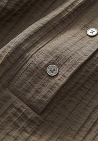 Tissu texturé brun clair avec deux boutons cousus, montrant des coutures détaillées et une petite section à rabat.