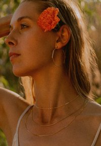 Goldene Creolen und geschichtete goldene Halsketten ergänzen das Subjekt, das eine leuchtend orange Blume im Haar und sonnenverwöhnte Haut hat.