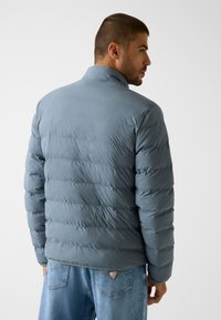 Szara pikowana kurtka z wysokim kołnierzem, z wypchanymi sekcjami i gładką fakturą, zestawiona z jasnoniebieskimi jeansami.