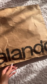 Rjava kraft papirnato vrečko z gubami, na sprednji strani je izrazito črna beseda "zalando". Vidna je roka, ki drži vrečko pred teksturirano ozadjem.
