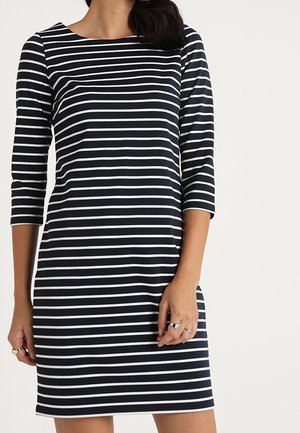 Femme portant une robe à rayures horizontales noires et blanches aux manches trois-quarts, debout devant un fond clair uni.