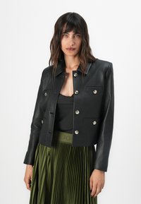 Veste en cuir noir coupée avec un col, boutons argentés et deux poches avant. Modèle portant une jupe plissée verte.