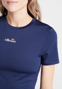 Námořně modré krátké tričko s krátkým rukávem, které má kulatý výstřih a malé vyšívané logo na horní části hrudi. Hladká textilie.