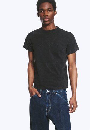 Jeune homme portant un t-shirt noir à manches courtes et un jean en denim bleu, une main dans la poche, debout devant un fond blanc uni.