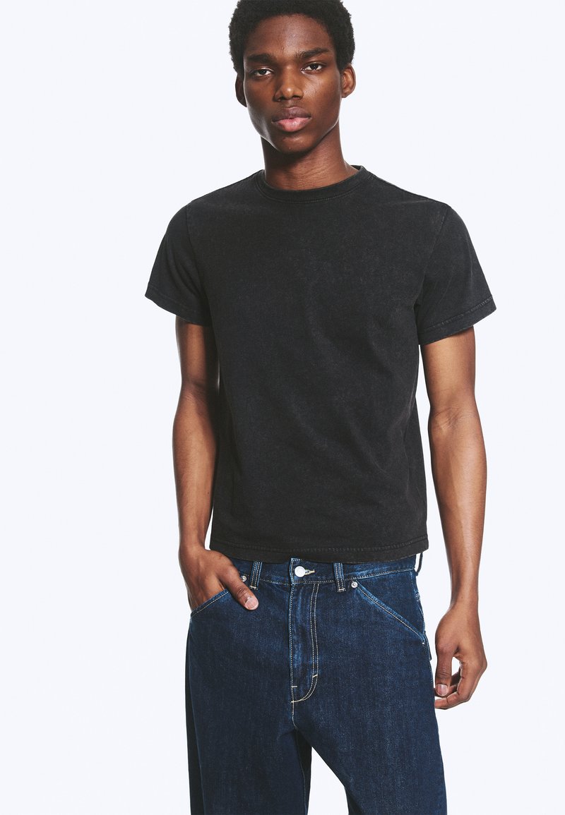 Jeune homme portant un t-shirt noir à manches courtes et un jean en denim bleu, une main dans la poche, debout devant un fond blanc uni.
