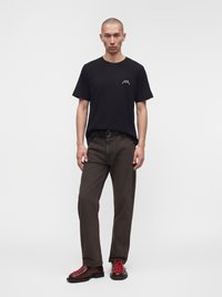 Homme portant un t-shirt noir à manches courtes, un pantalon marron foncé avec une ceinture noire et des bottes rouge foncé avec des lacets rouge vif, debout devant un fond uni.