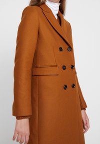 Double-breasted brown coat made of textured fabric, featuring black buttons, tailored lapels, and a side pocket. Styled with a white turtleneck.