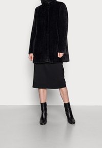 Sort faux fur frakke med høj krave, parret med en sort knælang nederdel og sorte ankelstøvler, sat mod en neutral baggrund.