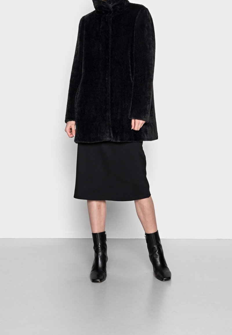 Sort faux fur frakke med høj krave, parret med en sort knælang nederdel og sorte ankelstøvler, sat mod en neutral baggrund.