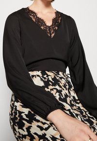 Blusa negra con escote en V y un intrincado borde de encaje negro. Las mangas largas y abullonadas, junto con una textura suave, se combinan con una falda estampada en beige y negro.