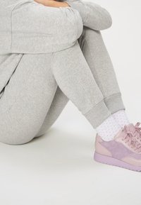 Personne assise, genoux ramenés, portant un pantalon de survêtement gris clair, des chaussettes blanches à petits pois, et des baskets roses sur un sol blanc.