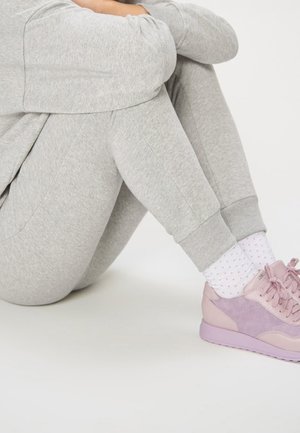 Osoba sedící se sepnutýma koleny, oblečená v světle šedých teplácích, bílých ponožkách s malými puntíky a růžových teniskách na bílém podlaze.