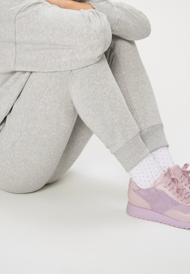 Personne assise, genoux ramenés, portant un pantalon de survêtement gris clair, des chaussettes blanches à petits pois, et des baskets roses sur un sol blanc.