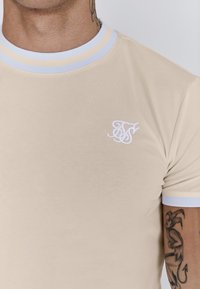 Beige T-shirt met een geribbelde lichtblauwe kraag, voorzien van een klein wit geborduurd logo aan de linkerkant. Soepele textuur.