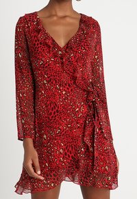 Robe portefeuille rouge avec un motif léopard, un col à volants, des manches longues et un ourlet flottant. Fabriquée en tissu léger et transparent.