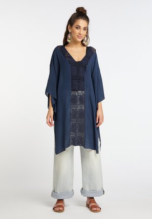 Frau trägt eine marineblaue Häkel-Tunika über hochgekrempelten hellen Jeans und braunen Sandalen, mit bunten großen Ohrringen und zu einem Dutt gebundenem Haar.
