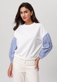 Vit sweatshirt med blå randiga puffärmar, ribbat nacke och midjeband. Kombinerad med ljusbeiga byxor.