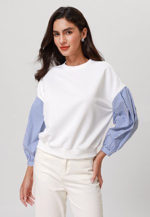 White sweatshirt with blue striped puff sleeves, ribbed neckline, and waistband. Paired with light beige pants.