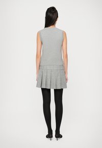 Top grigio senza maniche con maglia strutturata, abbinato a una gonna grigia plissettata. Il modello indossa collant neri e scarpe con tacco. Visto da dietro.