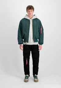 Zelená bomber bunda so zlatým zipsom, nosená cez šedý hoodie. V kombinácii s čiernymi cargo nohavicami s vreckami a červenou nášivkou. Zelené topánky.