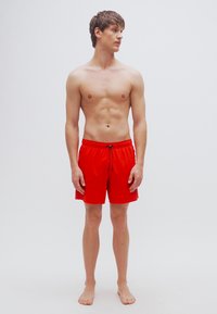 Κόκκινα μαγιό σορτς φτιαγμένα από ελαφρύ ύφασμα, με ελαστική μέση και κορδόνι. Το μοντέλο στέκεται ξυπόλητο με ουδέτερη στάση.