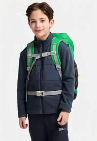 Groene rugzak met verstelbare riemen, grijze hardware en ademend gaas. Gedragen over een marineblauw fleecejack, met een gespsluiting aan de voorkant.