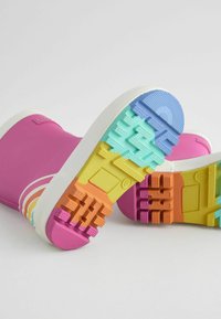 Bottes en caoutchouc roses avec une semelle blanche, présentant un profil de semelle à motifs multicolores en bleu, vert, jaune, orange et rose.