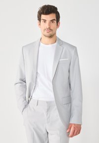 Hellgrauer Blazer mit taillierter Passform, zwei Knöpfen und einer Brusttasche. Getragen über einem weißen T-Shirt und kombiniert mit passenden Hosen.