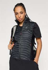 Černá izolační vesta s vysokým límcem, prošívanou strukturou, kapsami na zip a růžovými akcenty. Nosená přes černé tričko a s batohem.