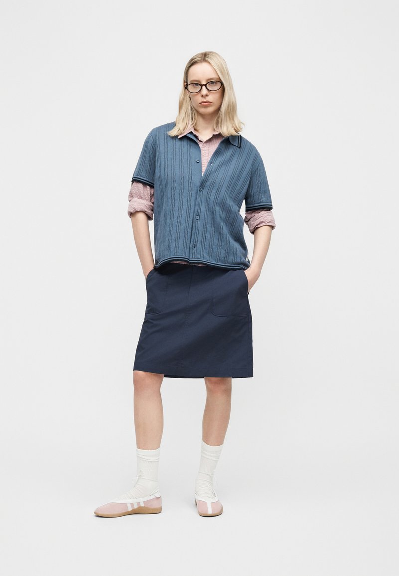 Donna bionda con occhiali, maglietta a maniche corte azzurra sopra una camicetta rosa con maniche arrotolate, gonna blu navy, calzini bianchi e scarpe rosa.