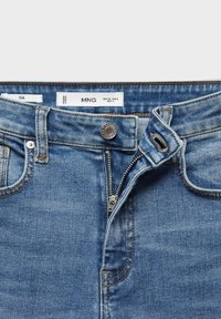 Jean en denim bleu avec fermeture éclair partiellement ouverte, bouton métallique, et étiquette de marque visible indiquant la taille et l'origine.