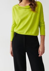 Top a maniche lunghe verde lime con scollatura a barca, abbinato a pantaloni neri su misura. Il tessuto appare morbido e leggero, con una vestibilità rilassata.