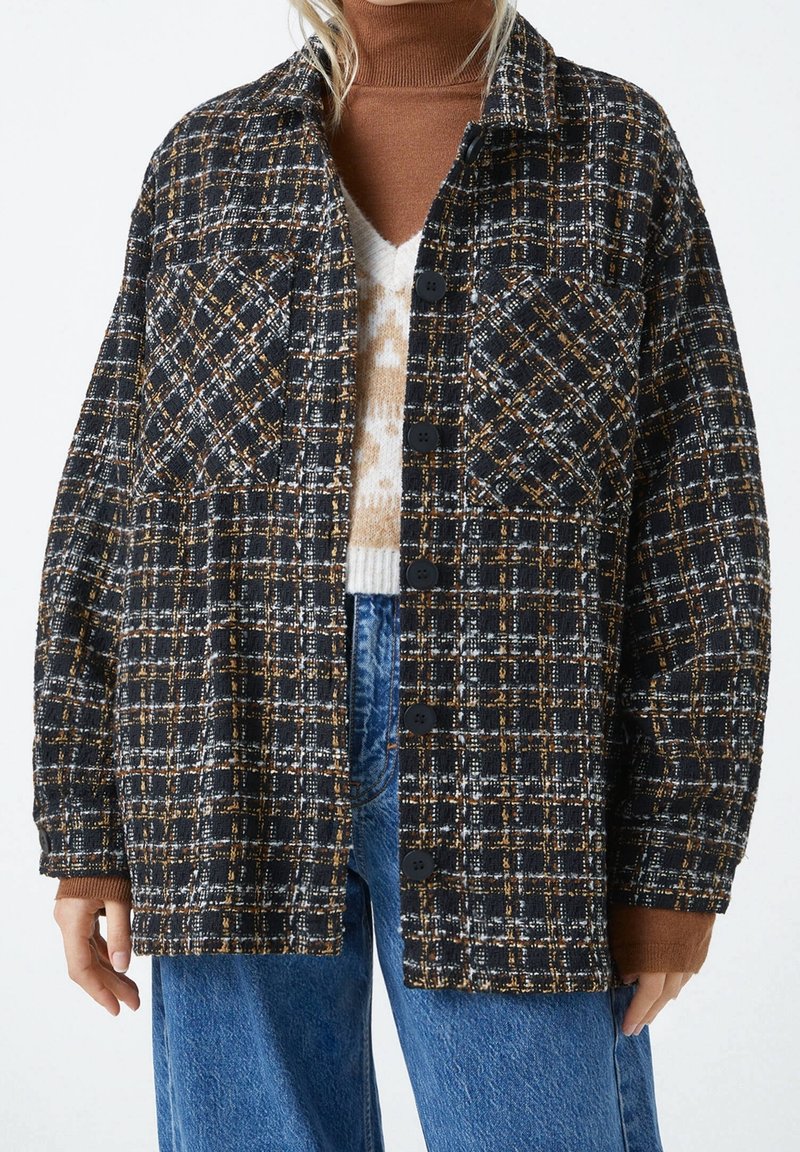 Personne portant une veste à carreaux noire et marron à boutons sur un col roulé marron et un jean bleu, les mains détendues le long du corps.